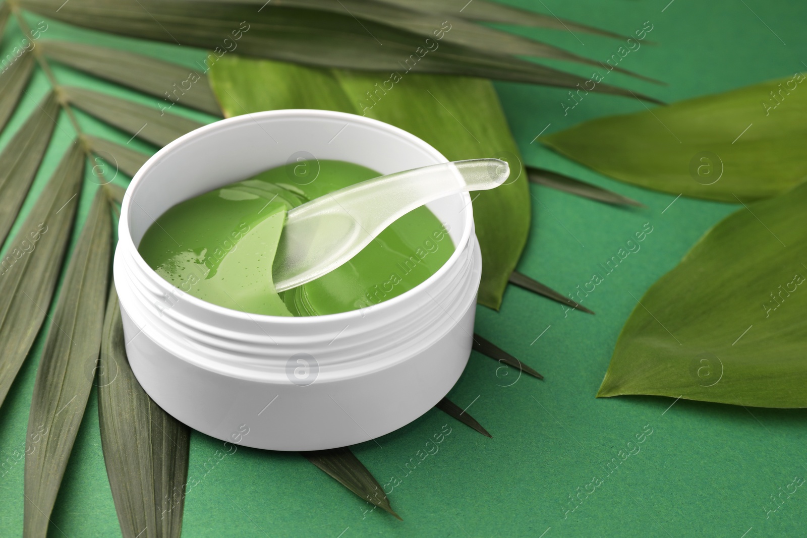 Photo of Jar of under eye patches with spoon and leaves on green background, closeup. Cosmetic product