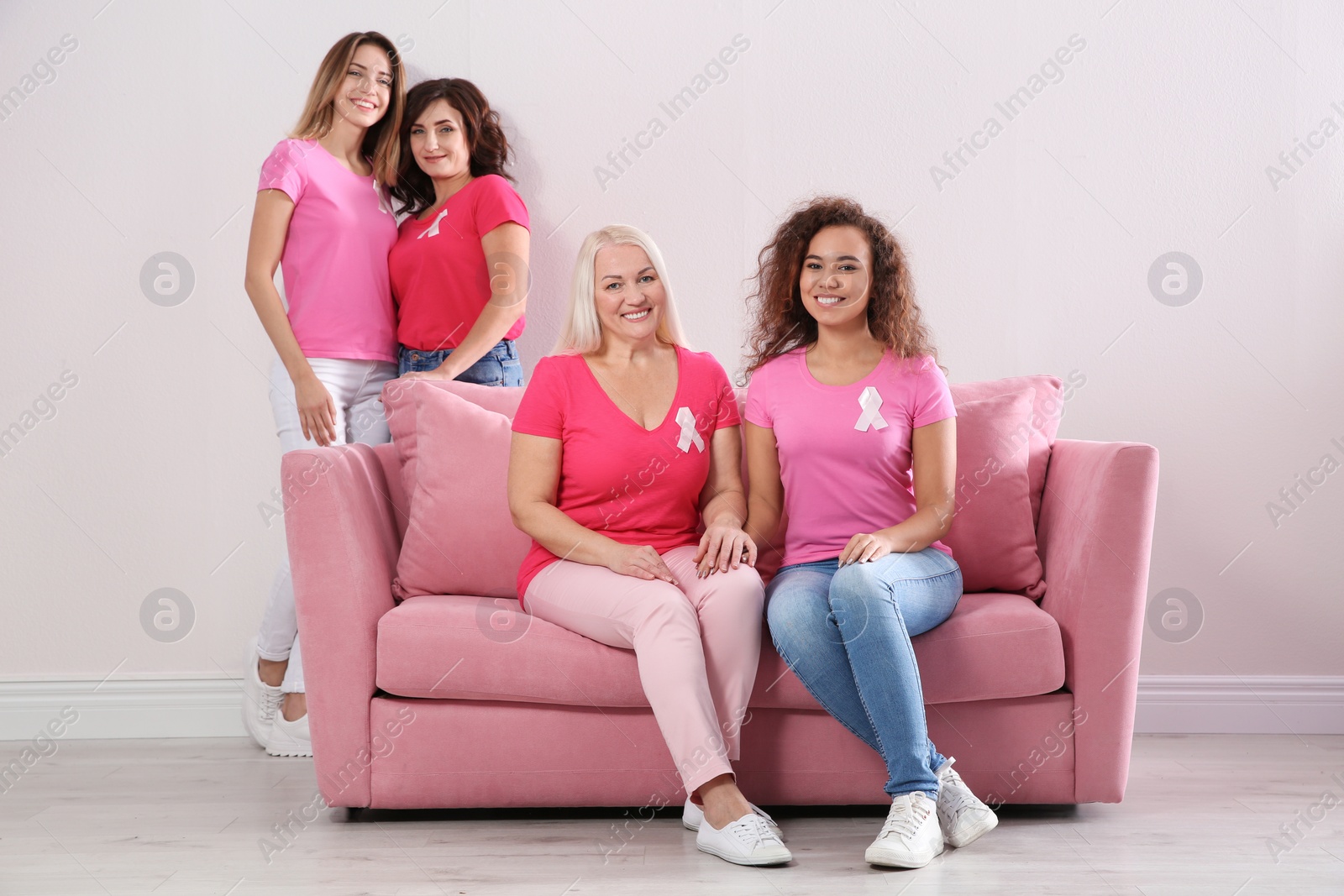 Photo of Group of women with silk ribbons sitting on sofa against light wall. Breast cancer awareness concept