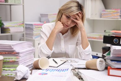 Photo of Overwhelmed woman surrounded by documents and paper coffee cups at workplace in office