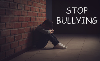 Image of Message STOP BULLYING and sad little boy sitting on floor near brick wall indoors