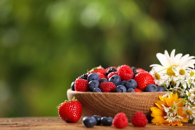 Photo of Bowl with different fresh ripe berries and beautiful flowers on table outdoors, space for text