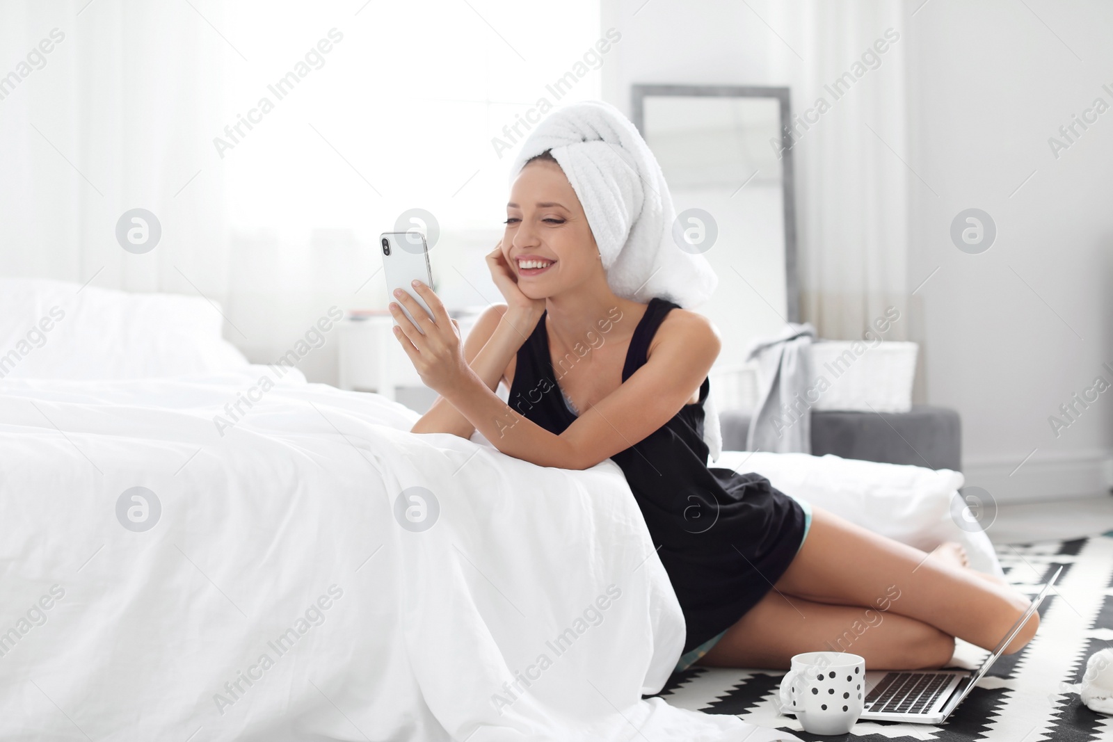 Photo of Young happy woman with mobile phone sitting on floor near bed at home. Lazy morning