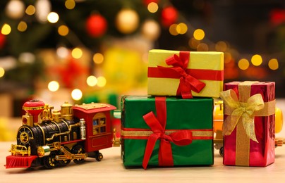 Beautiful gift boxes with toy train on wooden table against blurred festive lights. Christmas celebration