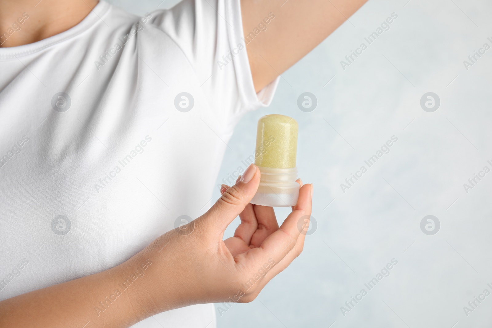 Photo of Young woman holding natural crystal alum deodorant near armpit on light blue background, closeup