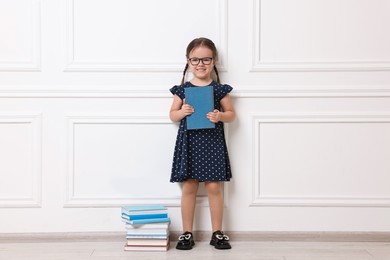 Photo of Cute little girl in glasses and stack of books near white wall