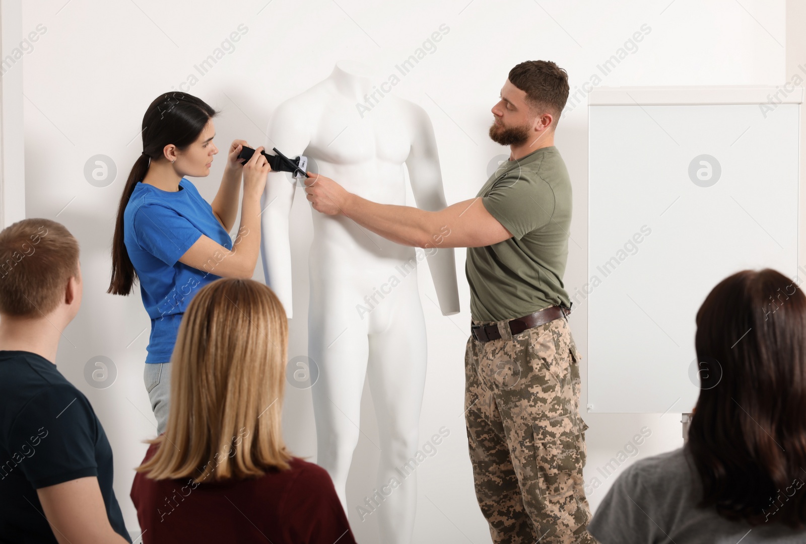 Photo of Soldier in military uniform teaching group of people how to apply medical tourniquet on mannequin indoors