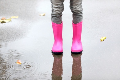 Photo of Little girl wearing rubber boots standing in puddle on rainy day, focus of legs. Autumn walk