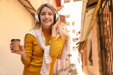 Photo of Happy young woman with coffee and headphones listening to music on city street
