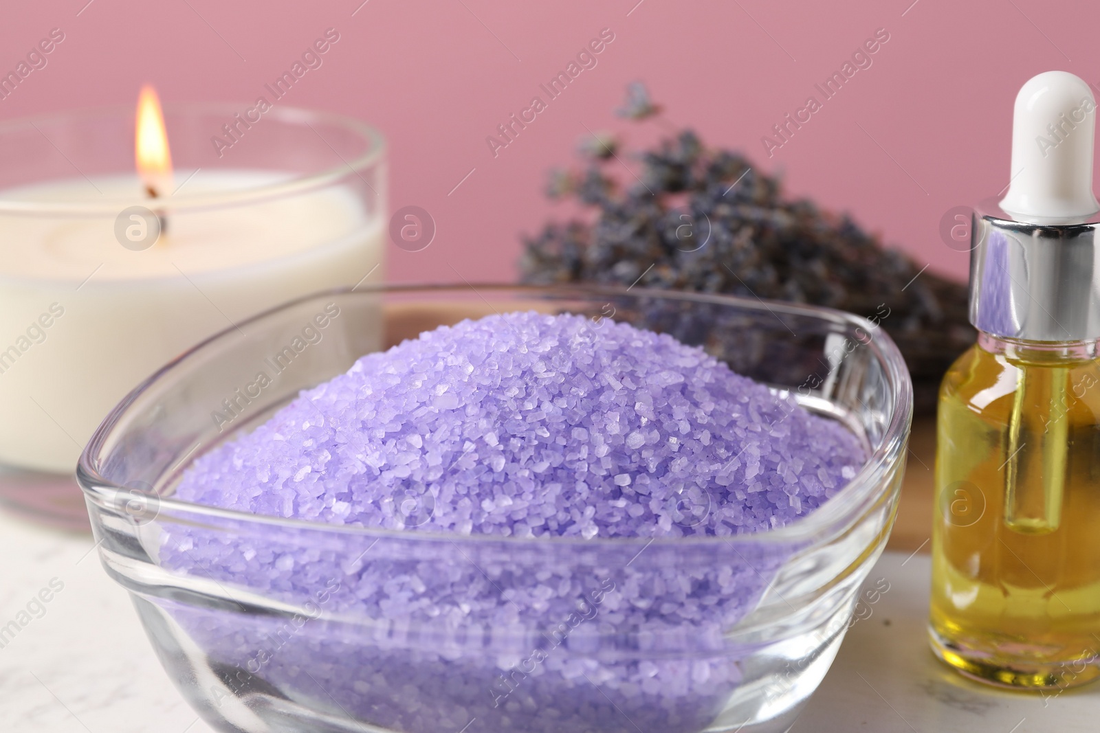 Photo of Violet sea salt in bowl, burning candle and cosmetic product on table, closeup