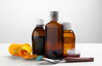 Photo of Bottles of syrup, measuring cup, lozenges and spoon on white wooden table against light grey background. Cold medicine