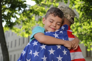 Photo of Soldier with flag of USA and his little son hugging outdoors