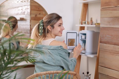 Photo of Woman taking cosmetic product from mini fridge indoors
