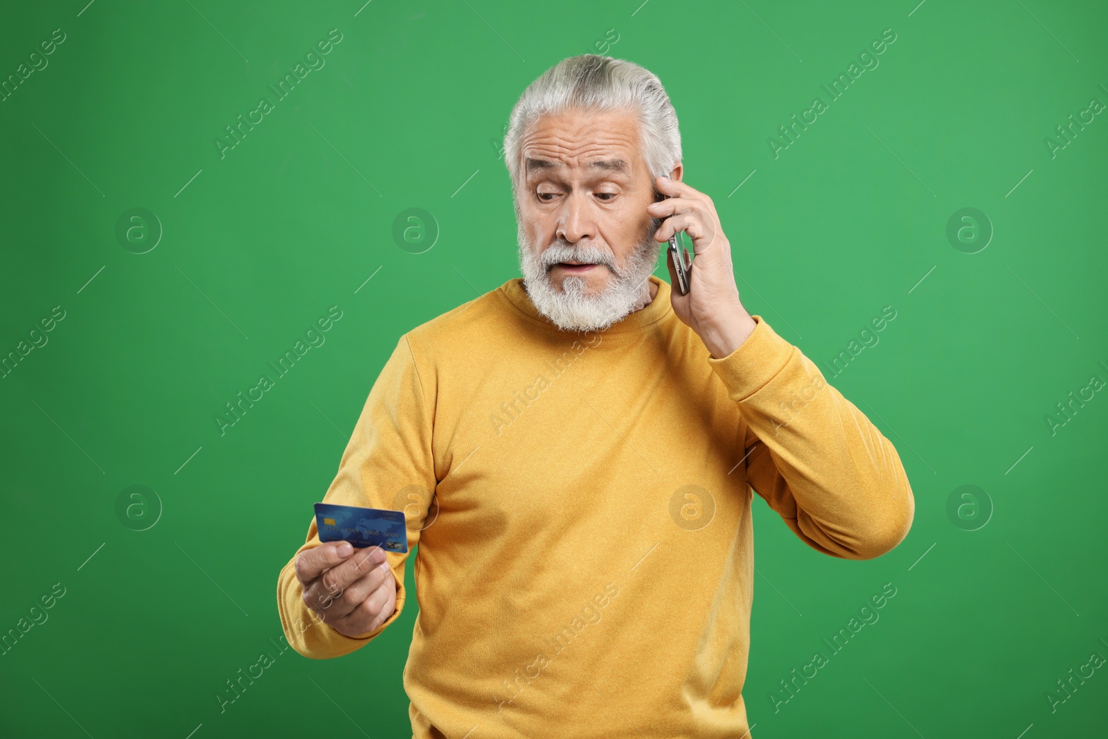 Photo of Confused senior man with credit card talking on smartphone against green background. Be careful - fraud