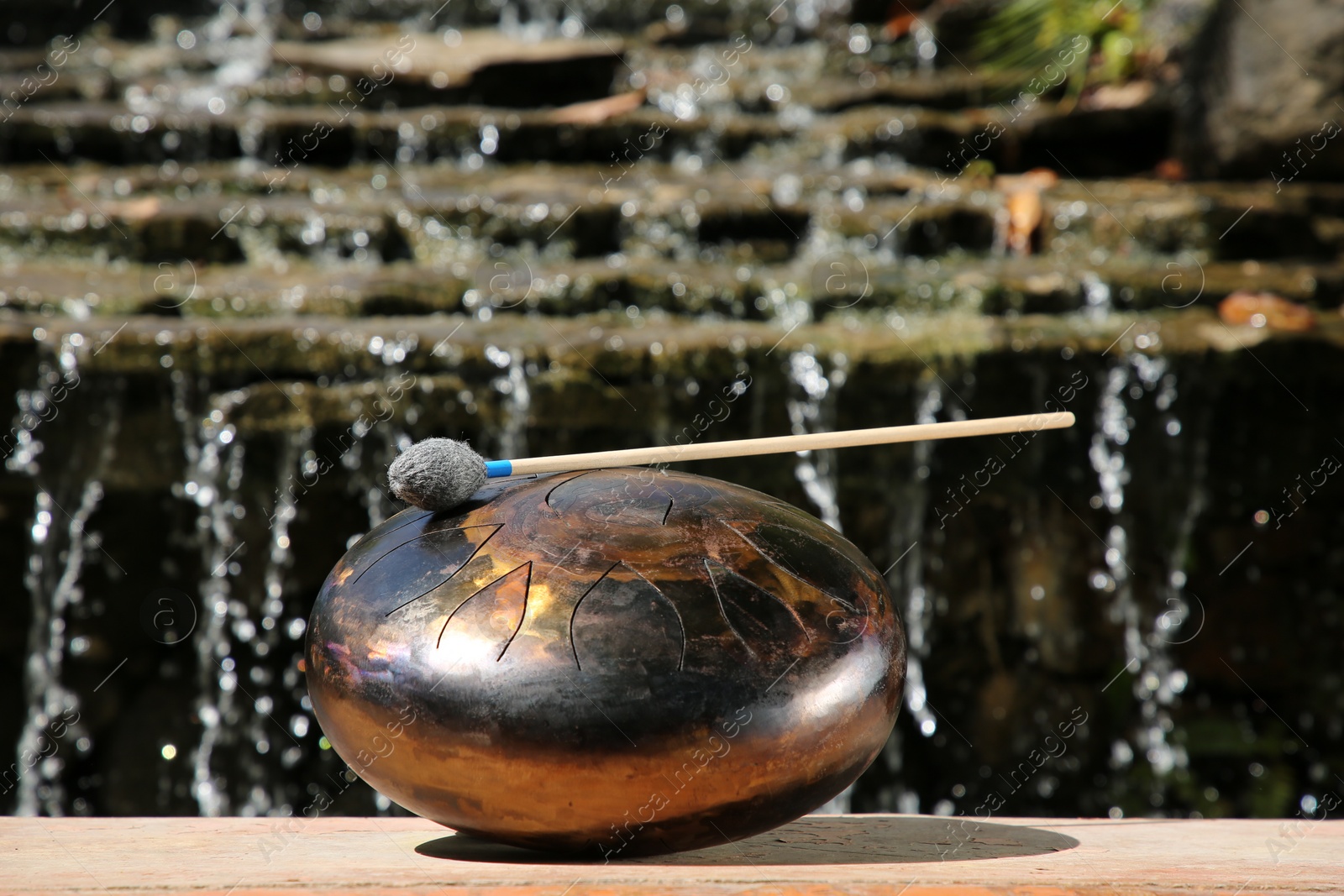 Photo of Steel tongue drum with mallet near waterfall outdoors on sunny day. Percussion musical instrument