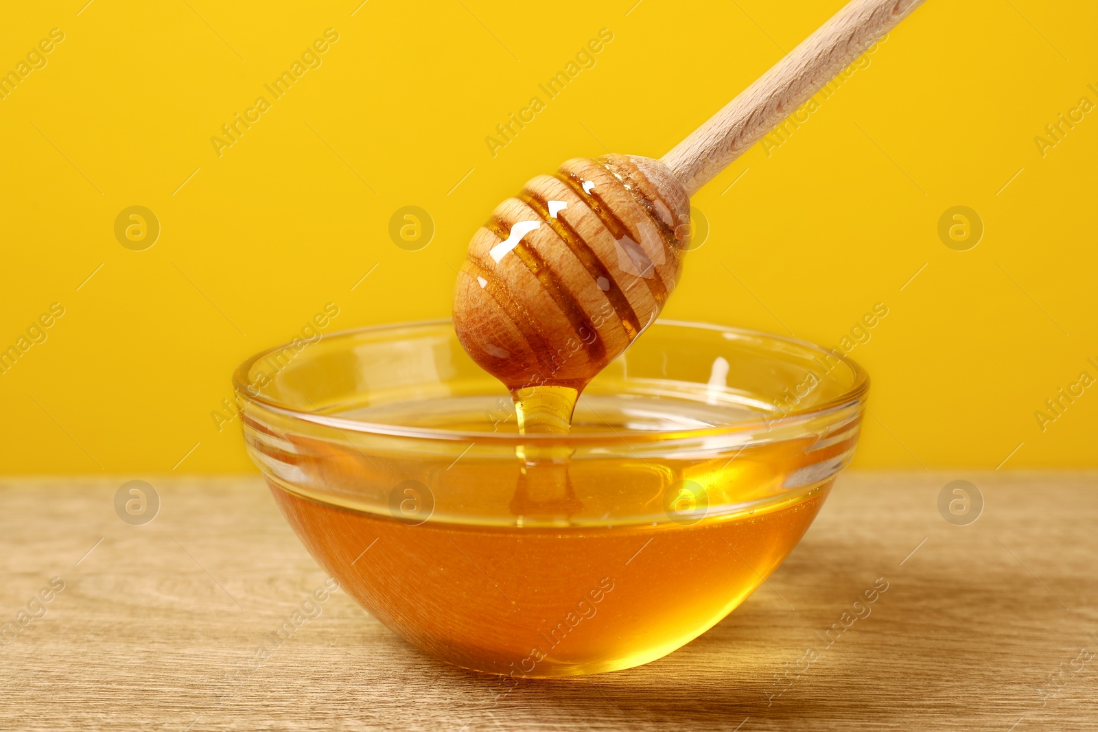 Photo of Pouring honey from dipper into glass bowl on wooden table against yellow background, closeup