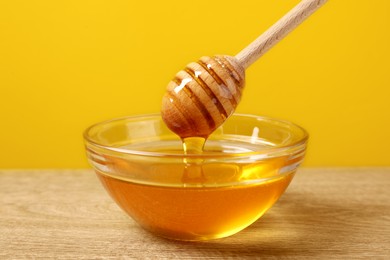 Photo of Pouring honey from dipper into glass bowl on wooden table against yellow background, closeup