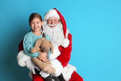 Photo of Little girl with toy bunny sitting on authentic Santa Claus' lap against color background