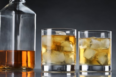 Tasty whiskey with ice in glasses and bottle on mirror table against dark background, closeup