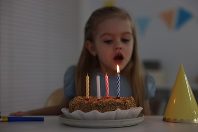 Photo of Cute girl blowing out birthday candles at table indoors, focus on cake