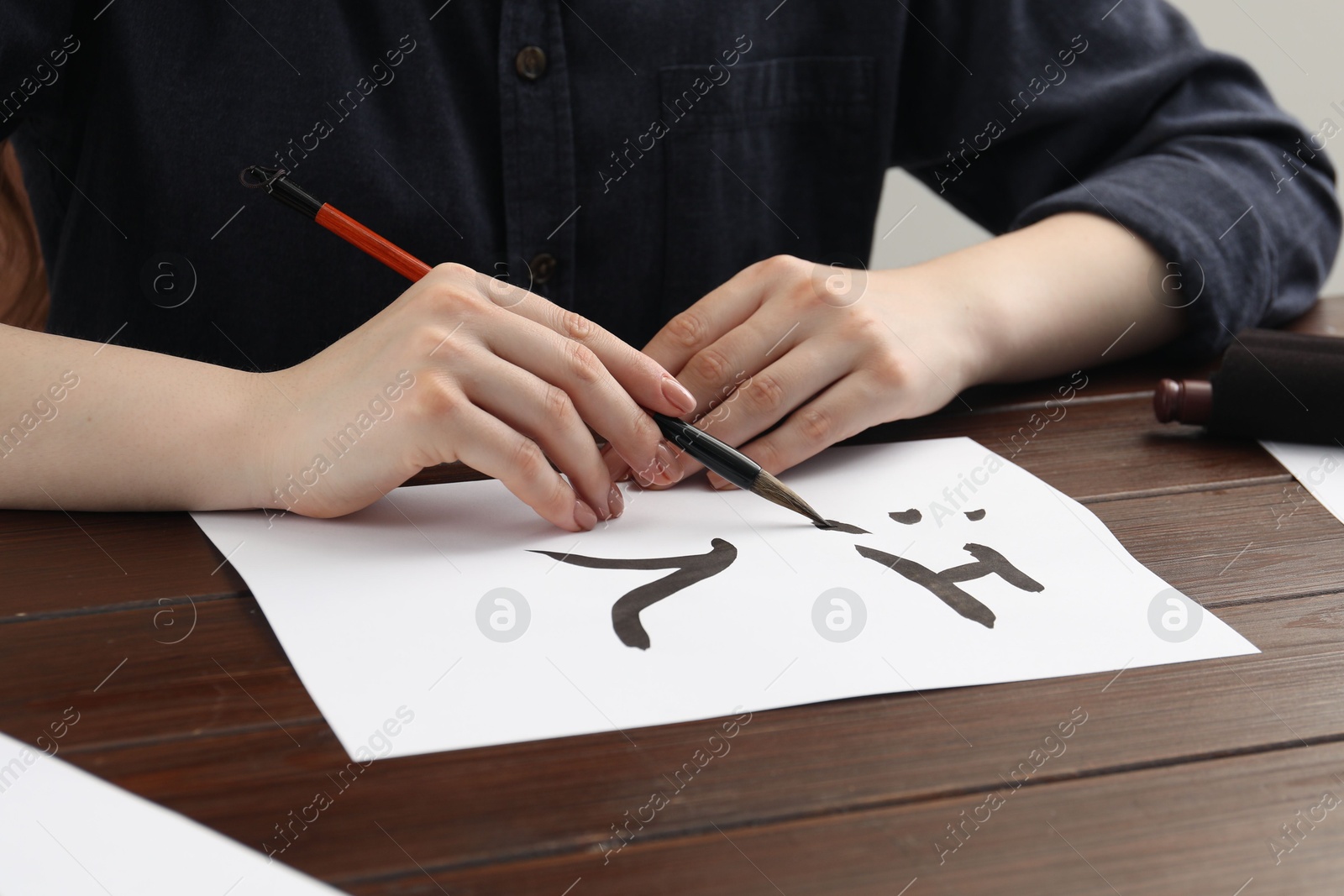 Photo of Calligraphy. Woman with brush writing hieroglyphs on paper at wooden table, closeup