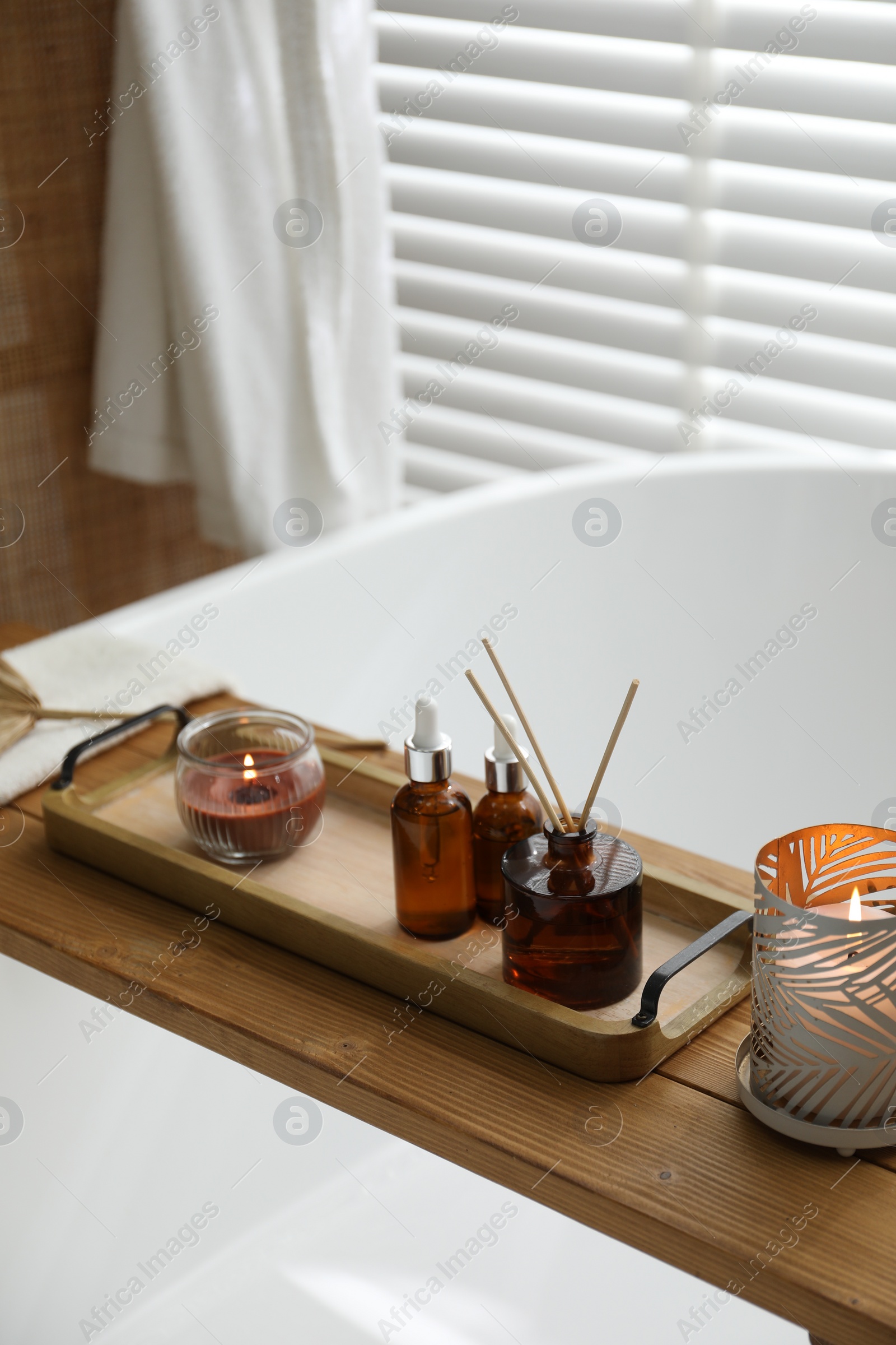 Photo of Wooden tray with cosmetic products, reed air freshener and burning candles on bath tub in bathroom