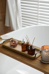 Wooden tray with cosmetic products, reed air freshener and burning candles on bath tub in bathroom