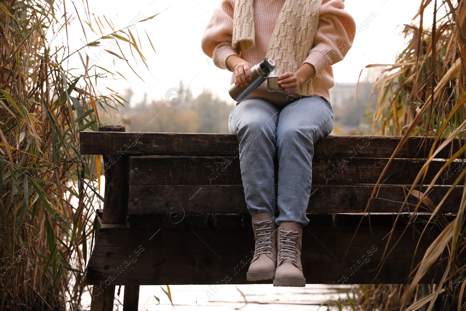 Photo of Woman with thermos sitting on wooden pier among reeds, closeup