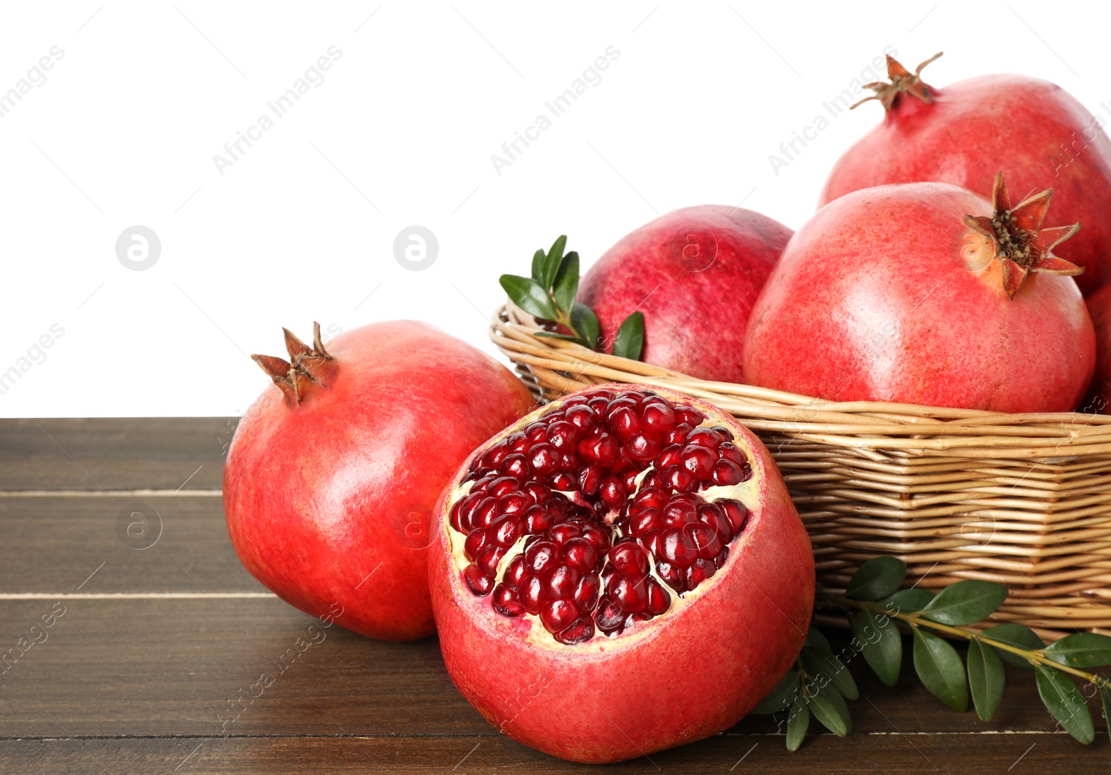 Photo of Fresh pomegranates in wicker basket and green leaves on wooden table against white background