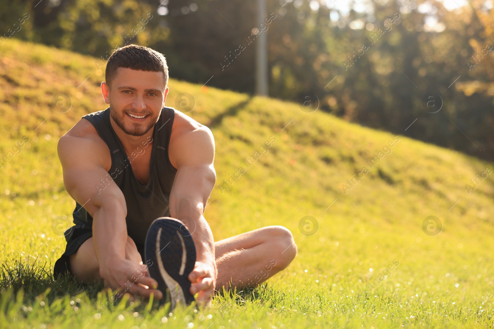 Photo of Attractive man doing exercises on green grass in park, space for text. Stretching outdoors