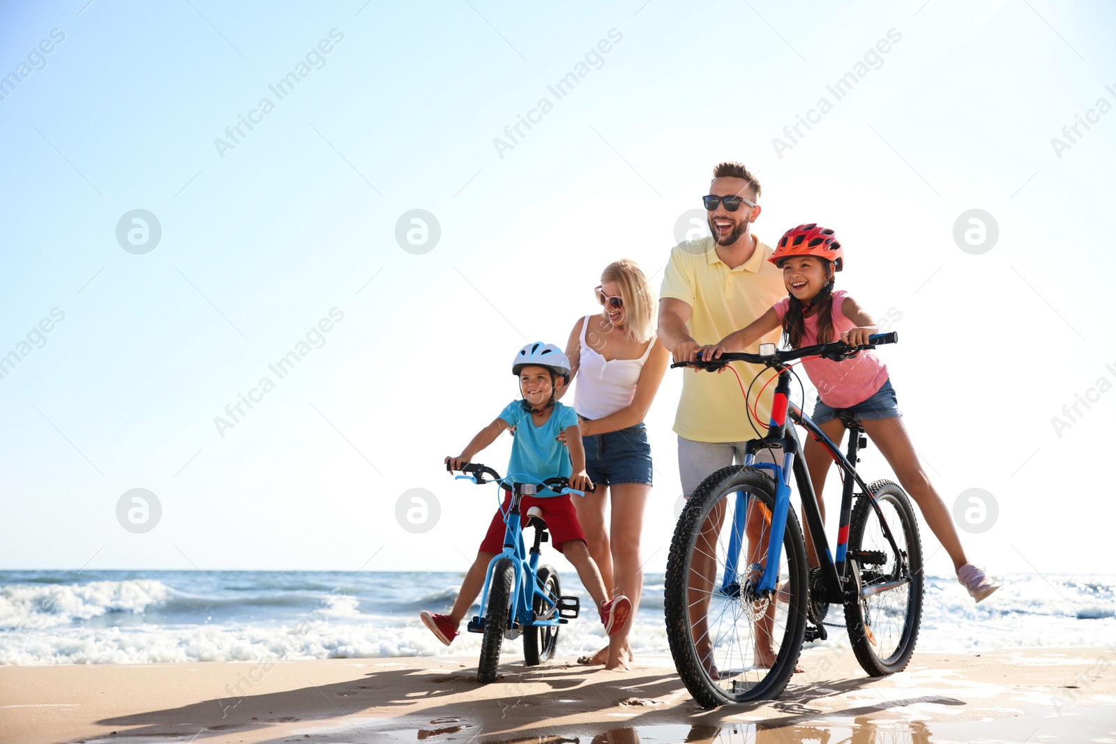 Photo of Happy parents teaching children to ride bicycles on sandy beach near sea