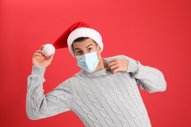 Emotional man wearing Santa hat and medical mask on red background