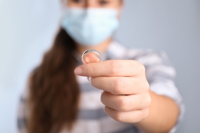 Photo of Woman in protective mask holding wedding ring against light background, focus on hand. Divorce during coronavirus quarantine