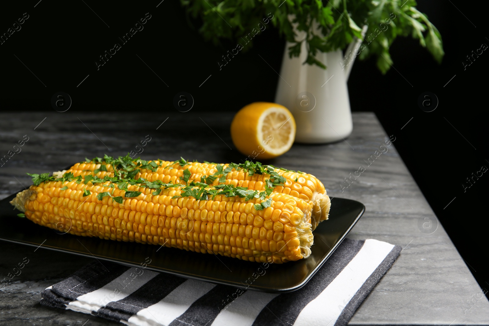 Photo of Delicious boiled corn cobs with parsley on gray table against black background