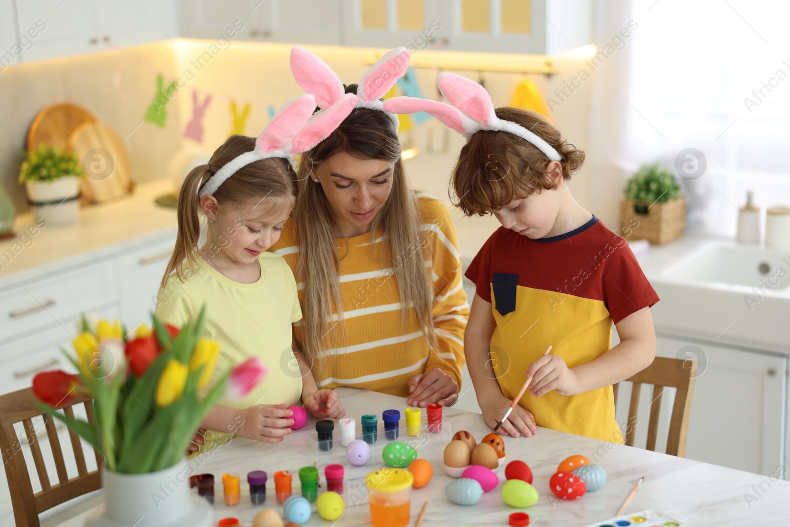 Photo of Easter celebration. Mother and her cute children with bunny ears painting eggs at white marble table in kitchen