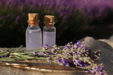 Photo of Bottles of natural lavender essential oil and flowers on burlap cloth outdoors