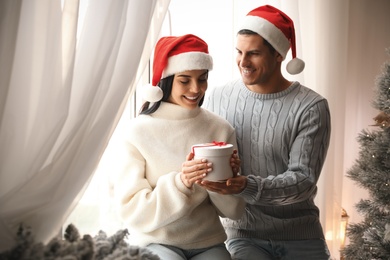 Photo of Couple in Santa hats holding gift box near Christmas tree