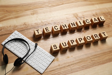 Photo of Cubes with words TECHNICAL SUPPORT, headset and computer keyboard on wooden background