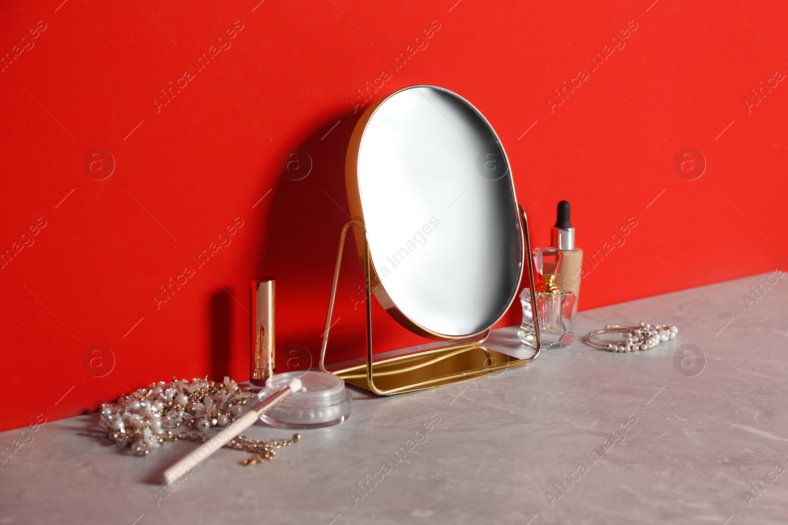 Photo of Small mirror, makeup products and jewelry on grey marble table near red wall