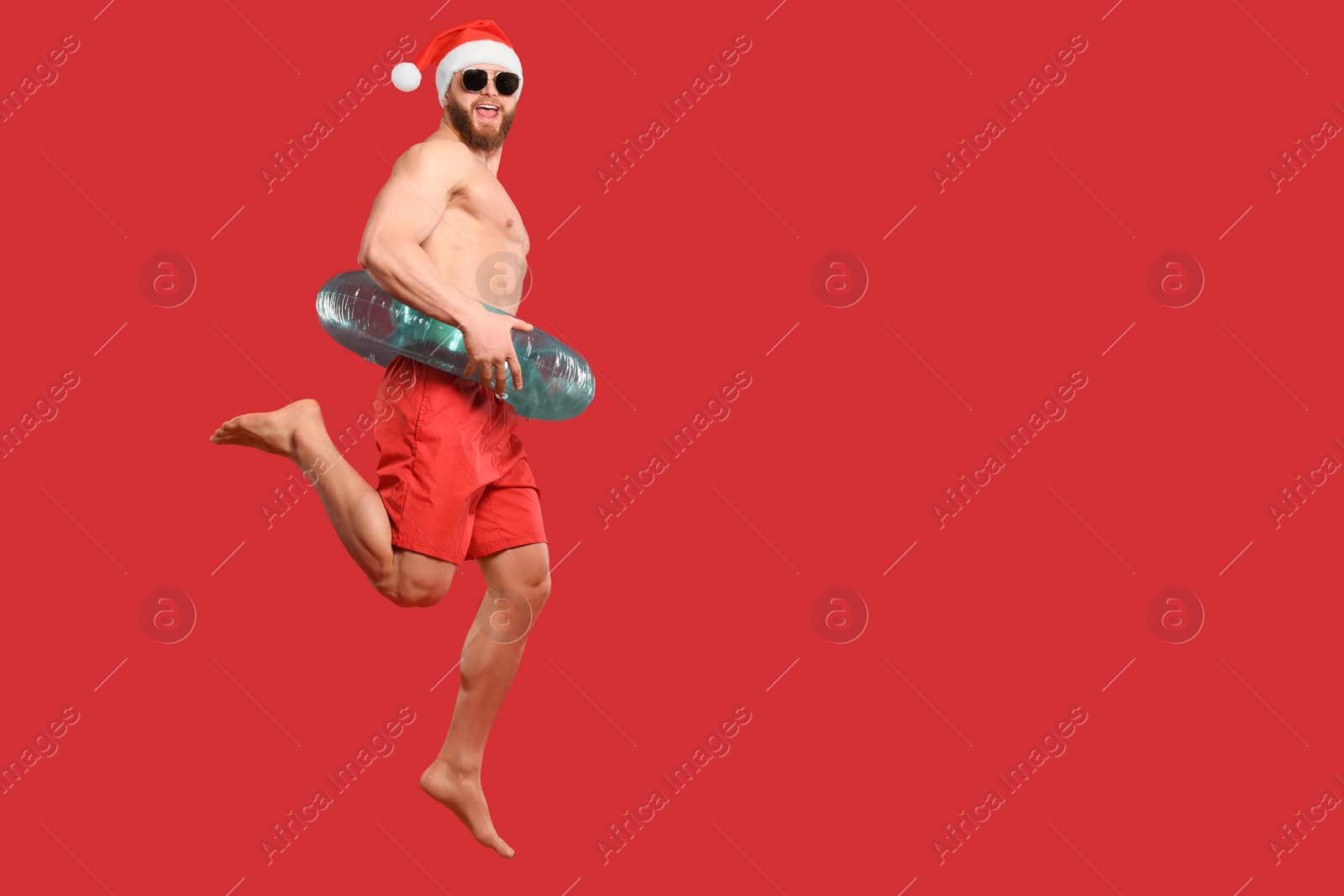 Photo of Muscular young man in Santa hat with inflatable ring jumping on red background, space for text