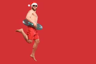 Photo of Muscular young man in Santa hat with inflatable ring jumping on red background, space for text