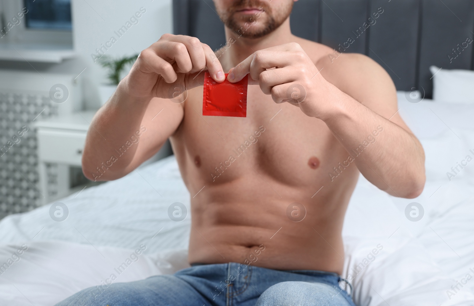 Photo of Closeup view of man opening pack with condom on bed indoors