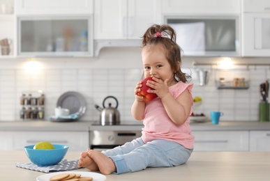 Adorable little baby girl eating apple on table in kitchen