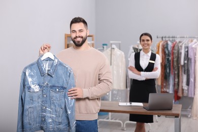 Photo of Dry-cleaning service. Man holding hanger with denim jacket indoors. Happy worker at workplace