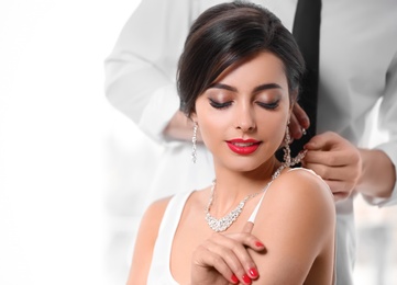 Man putting elegant jewelry on beautiful woman against light background. Space for text