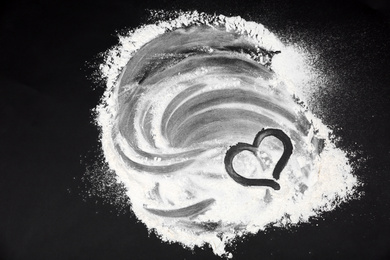 Photo of Drawing of heart on flour scattered over black table, top view