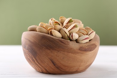 Photo of Tasty pistachios in bowl on white wooden table against olive background, closeup