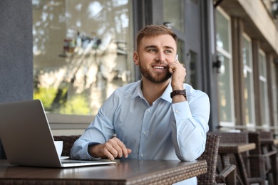 Image of Happy young man with laptop talking on phone at outdoor cafe