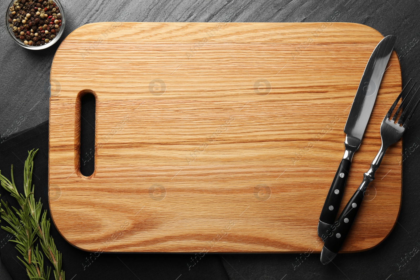 Photo of Wooden cutting board, spices and cutlery on dark grey table, flat lay