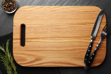 Wooden cutting board, spices and cutlery on dark grey table, flat lay