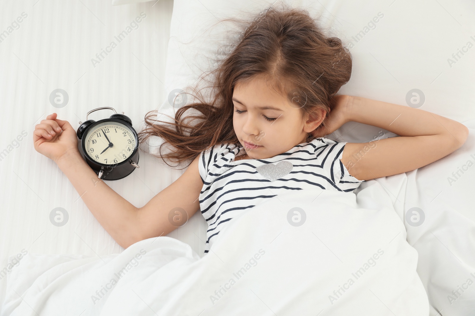 Photo of Cute little girl with alarm clock sleeping in bed, above view